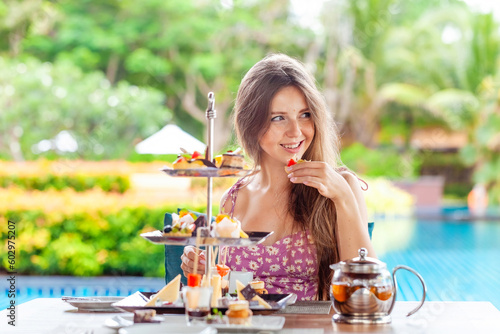 Young woman eat sweet tart cake, sits by table with afternoon tea set in outdoors poolside cafe or restaurant at luxury tropical resort or hotel with pool background. Summer holiday, vacation concept.