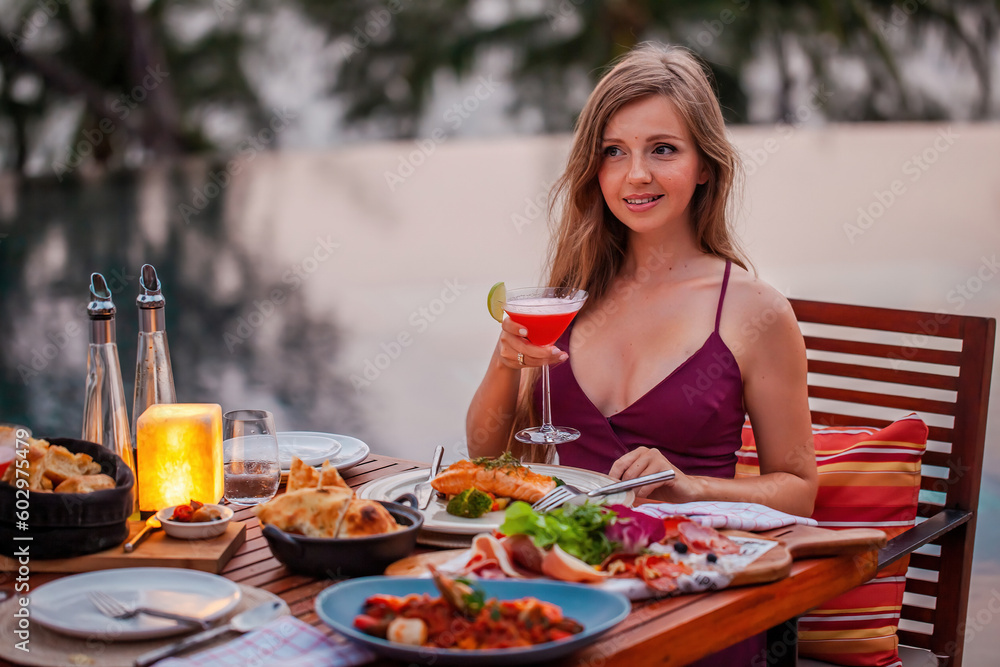 Elegant attractive smiling woman on romantic dinner in luxury gourmet restaurant. Happy young lady with a glass of cocktail in fine dining outdoor restaurant. Sexy elegant girl celebrating holiday. Elegant attractive smiling woman on romantic dinner in luxury gourmet restaurant. Happy young lady with a glass of cocktail in fine dining outdoor restaurant. Sexy elegant girl celebrating holiday.