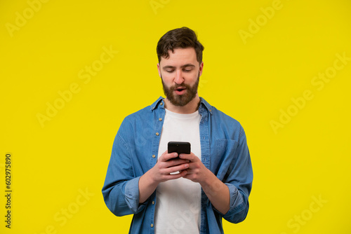 Bearded, cheerful, attractive guy in casual outfit, jeans shirt, holding smart phone in hands, using 3G internet, wi-fi, checking email, doing online shopping over yellow background