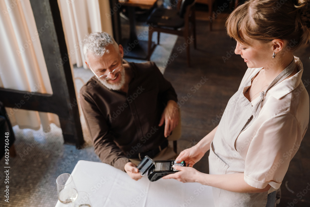Cheerful young waitress accepts payment through bank terminal for lunch of happy mature man in cafe Cheerful young waitress accepts payment through bank terminal for lunch of happy mature man in cafe