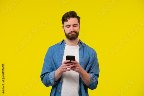 Bearded, cheerful, attractive guy in casual outfit, jeans shirt, holding smart phone in hands, using 3G internet, wi-fi, checking email, doing online shopping over yellow background © kinomaster