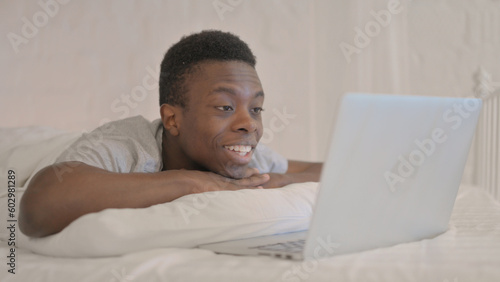 Young African Man Doing Video Chat on Laptop while Lying on Stomach in Bed