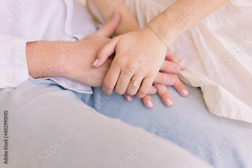 A tender, romantic moment of a romantic couple holding hands. He wears jeans and she wears a white dress, her hands with the engagement ring are on top of his.