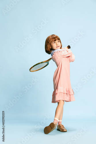 Full-length portrait of smiling, cute, little girl in pink dress posing with tennis racket against blue studio background. Concept of childhood, emotions, fun, fashion, active lifestyle, sport