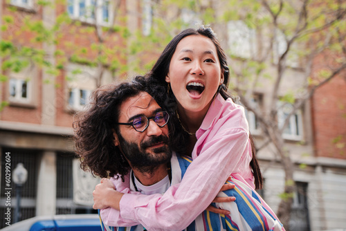 Young multiracial happy couple having fun embracing and hugging toguether. Caucasian bearded husband smiling and giving a piggyback ride to his asian lovely wife on a romantic date or honneymoon. High photo