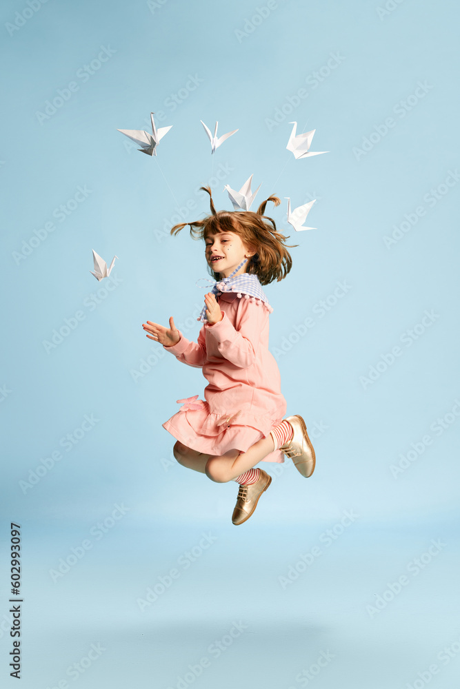 Full-length portrait of playful, lovely little girl in pink dress posing, playing with paper birds against blue studio background. Concept of childhood, emotions, fun, fashion, lifestyle, imagination Full-length portrait of playful, lovely little girl in pink dress posing, playing with paper birds against blue studio background. Concept of childhood, emotions, fun, fashion, lifestyle, imagination