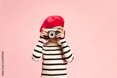 Portrait of little girl, cild in striped sweater and red beret taking photo with vintage photo camra agaisnt pink studio background. Concept of childhood, emotions, fun, fashion, lifestyle, travelling © master1305