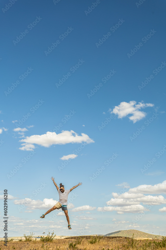 long haired man jumping with outstretched arms in a meadow with white clouds and blue sky in the background long haired man jumping with outstretched arms in a meadow with white clouds and blue sky in the background