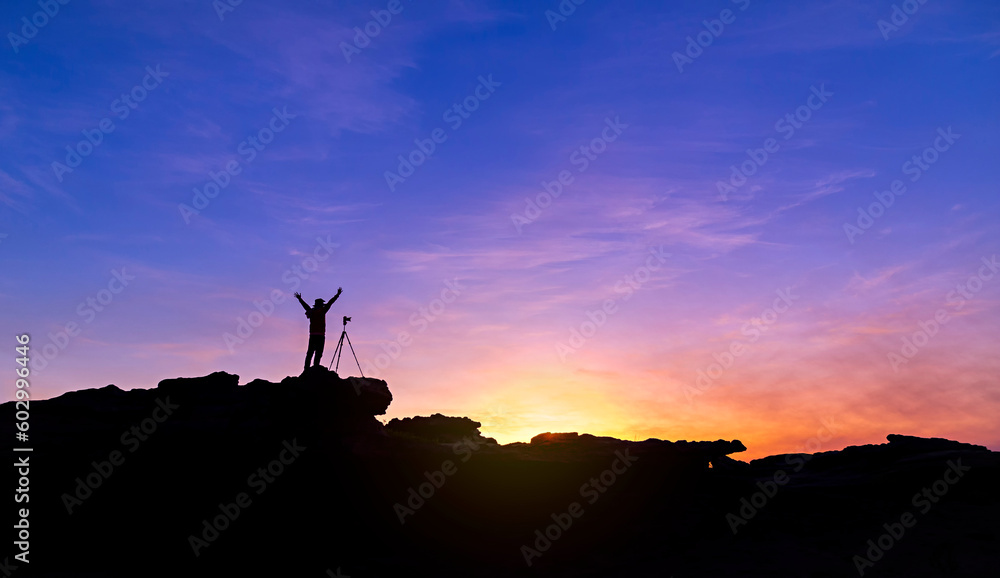 Silhouette of a man photographer standing on top of mountain at sunrise background. The photographer raised hands with the tripod takes photos high in the mountains.