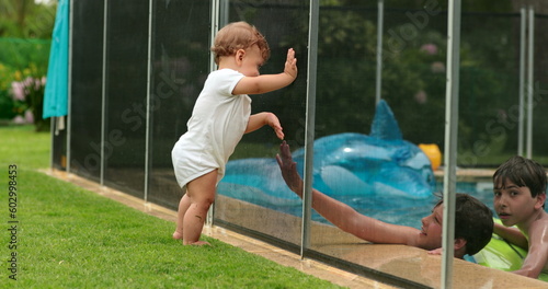 Baby looking at kids play inside swimming pool water during sumemr day. infant leaning on pool fence photo