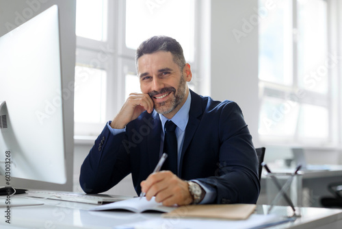 Happy middle aged businessman writing in notepad while working in office with computer, planning business schedule