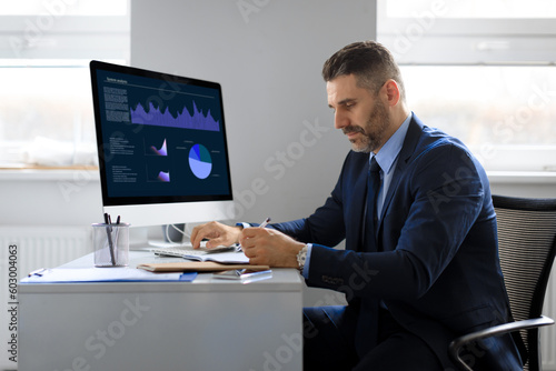 Middle aged businessman working on computer, sitting in front of monitor with online trading statistics in office