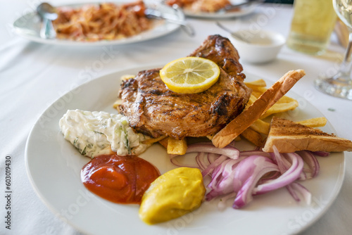 Roasted pork steak with French fries, onions, various dips and a lemon slice, barbecue dish in summer on a white plate, selected focus
