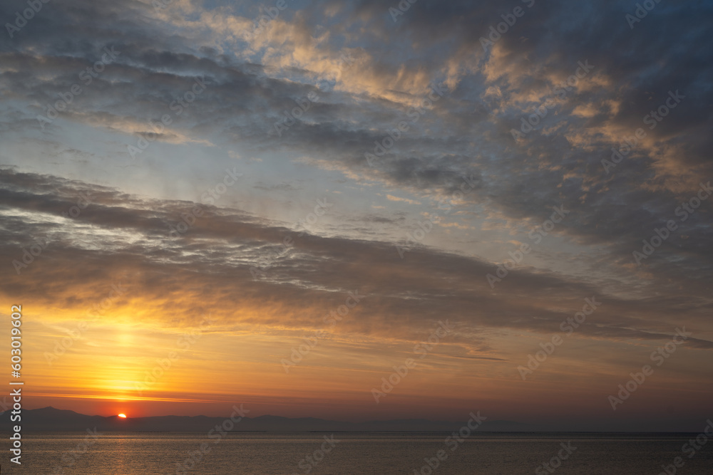 Red glowing sun half behind the mountains by the sea at sunset or sunrise illuminating the twilight sky with light stripes of clouds in orange to blue, romantic weather and climate concept, copy space