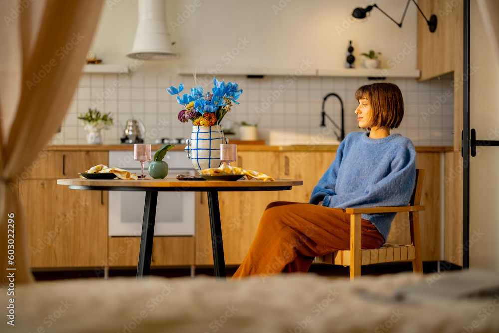 Young woman sitting relaxed by the dining table decorated with fresh flowers in modern and stylish studio apartment. Home coziness and style concept