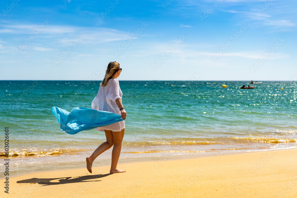 Beautiful woman walking on sunny beach holding shawl Beautiful woman walking on sunny beach holding shawl