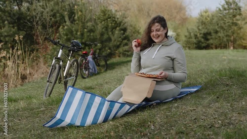 Plus size caucasian woman sits on grassy hillock in park, enjoys sniffing apple, wide shot. On lap lies pizza, woman chooses healthy diet. Wind ruffles hair, lifts rug. Two bicycles stand near person. photo