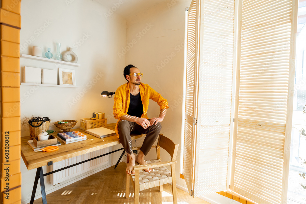 Portrait of creative man sitting on wooden table with books at workspace of stylish apartment. Hobby, creativity and home office concept Portrait of creative man sitting on wooden table with books at workspace of stylish apartment. Hobby, creativity and home office concept