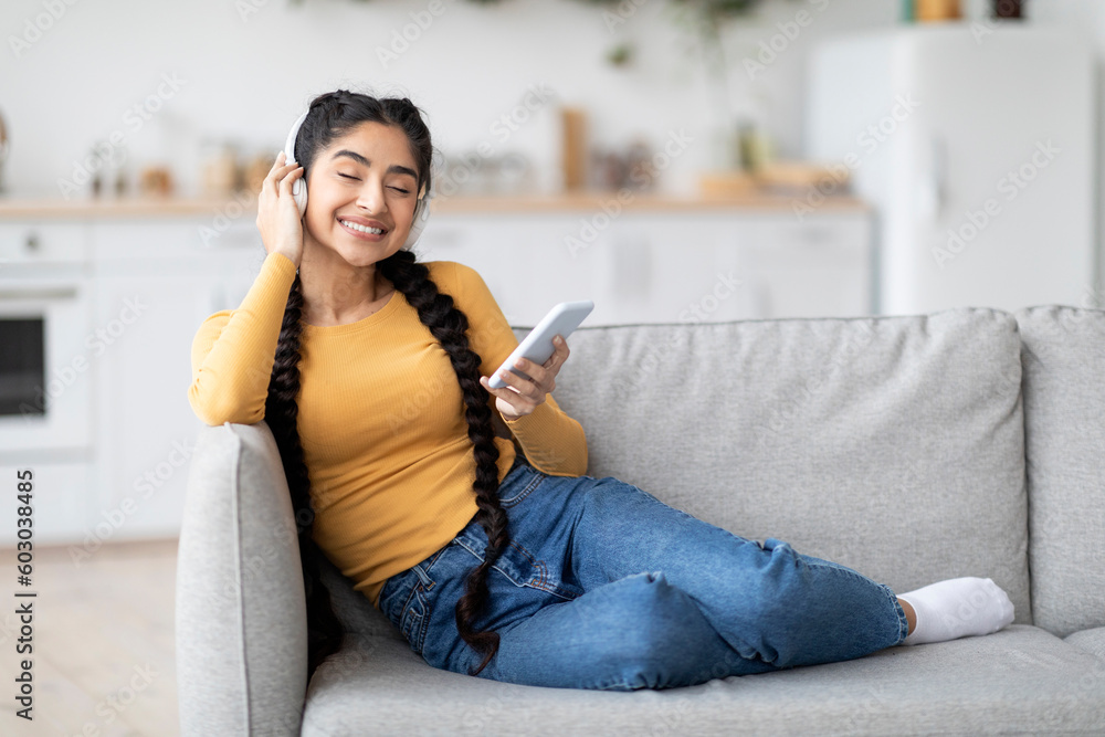 Obraz premium Happy Indian Female Wearing Wireless Headphones Relaxing With Smartphone At Home