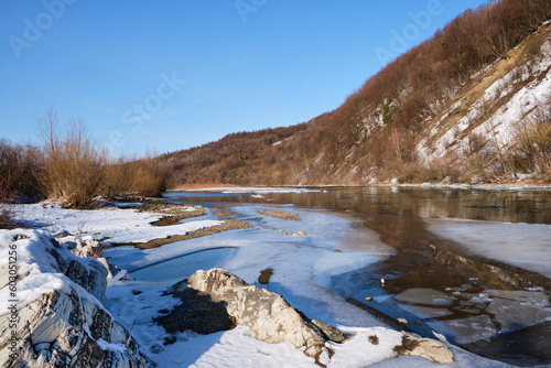 Fototapeta Naklejka Na Ścianę i Meble -  Winter landscape on a clear day. A view of a mountain river with ice and mountains covered with snow and stones with different structures and a clear sky