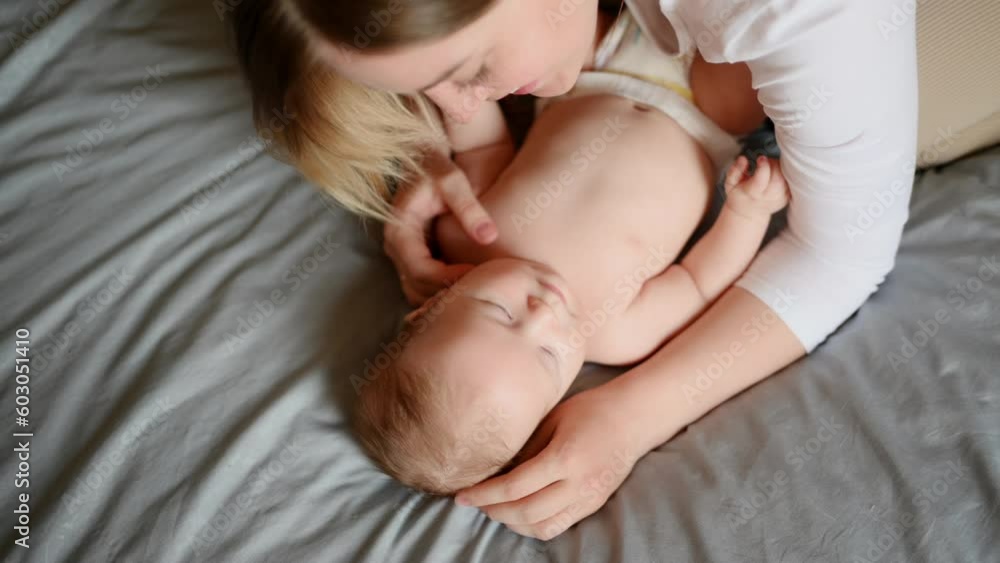 Joyful young mother plays with her baby lying on the bed, smiles, kisses the cheek, enjoys motherhood.Family, love,care. Joyful young mother plays with her baby lying on the bed, smiles, kisses the cheek, enjoys motherhood.Family, love,care.