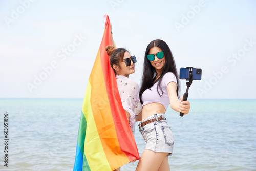 LGBT lesbian couple with a rainbow flag and taking selfie from smartphone on the beach