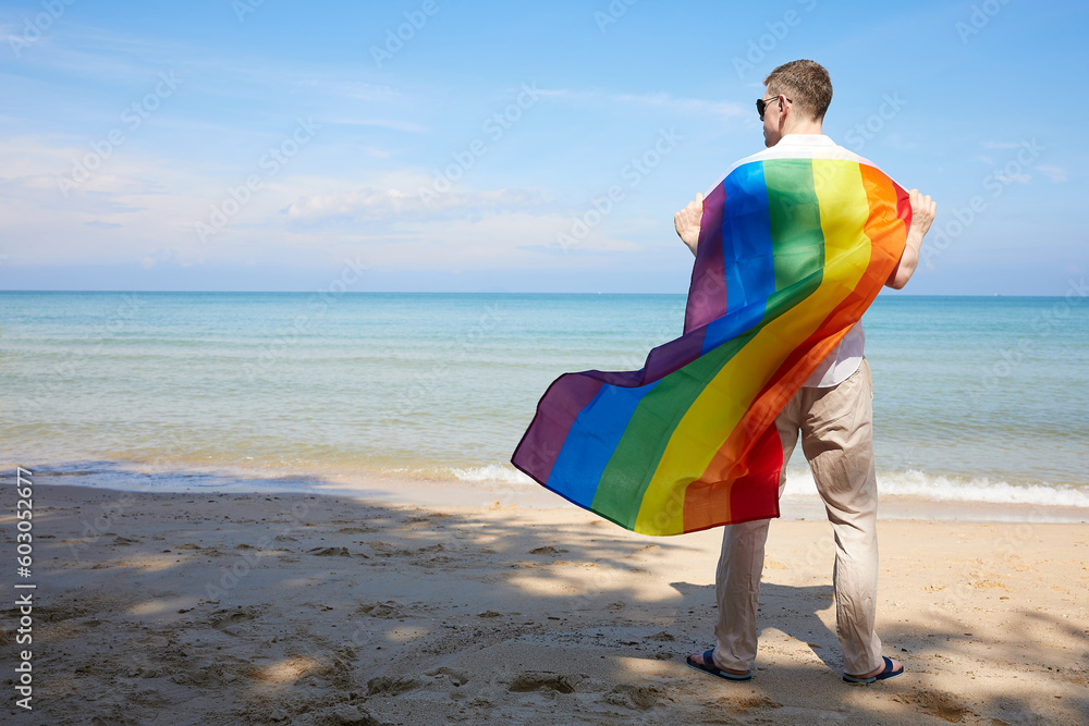 back young man looking to the sea and holding a rainbow flag(LGBT) on a tropical beach