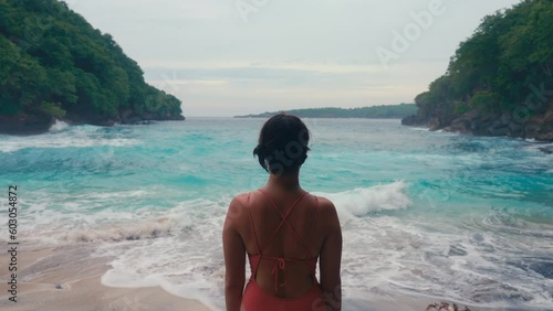 A lonely woman with short hair standing on wild ocean beach with turquoise water looking on waves. Pensive thoughtful person on seaside. Relaxation and enjoying nature on summer holidays. Middle plan