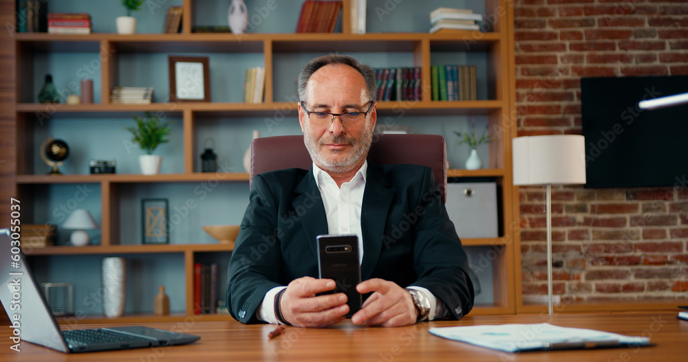 Joyful smiling middle age man chatting use smartphone enjoying break during work in front of laptop