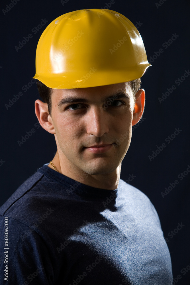 Handsome brunette young man wearing a yellow hard hat and navy blue tshirt standing on black background Handsome brunette young man wearing a yellow hard hat and navy blue tshirt standing on black background