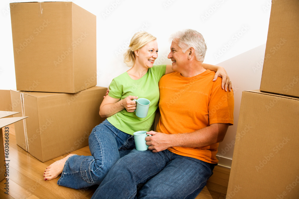 Middle-aged couple sitting on floor among cardboard moving boxes drinking coffee. Middle-aged couple sitting on floor among cardboard moving boxes drinking coffee.
