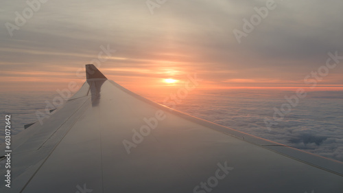View along jet aircraft wing with orange sun on horizon in flight above clouds