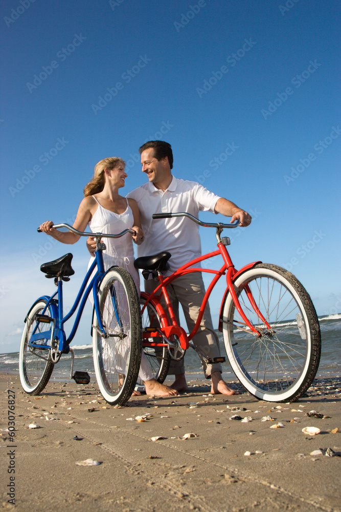 Man and woman look at each other while pushing bikes on the beach. Vertical shot.