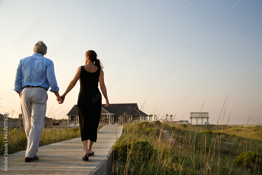 Fototapeta premium Couple walk hand in hand on a boardwalk towards a beach pavilion. Horizontal shot.