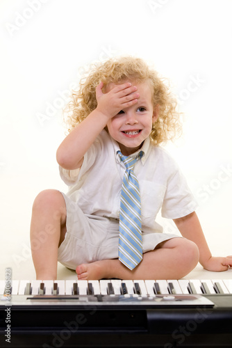 Adorable little three year old boy with long curly blond hair wearing white shirt and tie sitting in front of a piano keyboard with hand on forhead photo