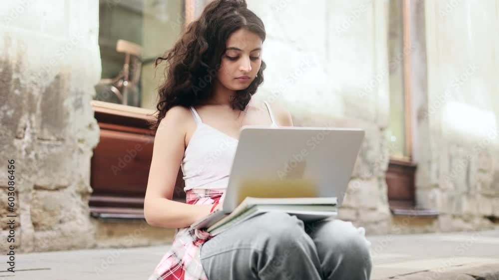 Pretty young indian businesswoman freelancer working on laptop sitting on border at city street Busy focused curly woman have distance remote freelance work on computer outdoors alone Pretty young indian businesswoman freelancer working on laptop sitting on border at city street Busy focused curly woman have distance remote freelance work on computer outdoors alone