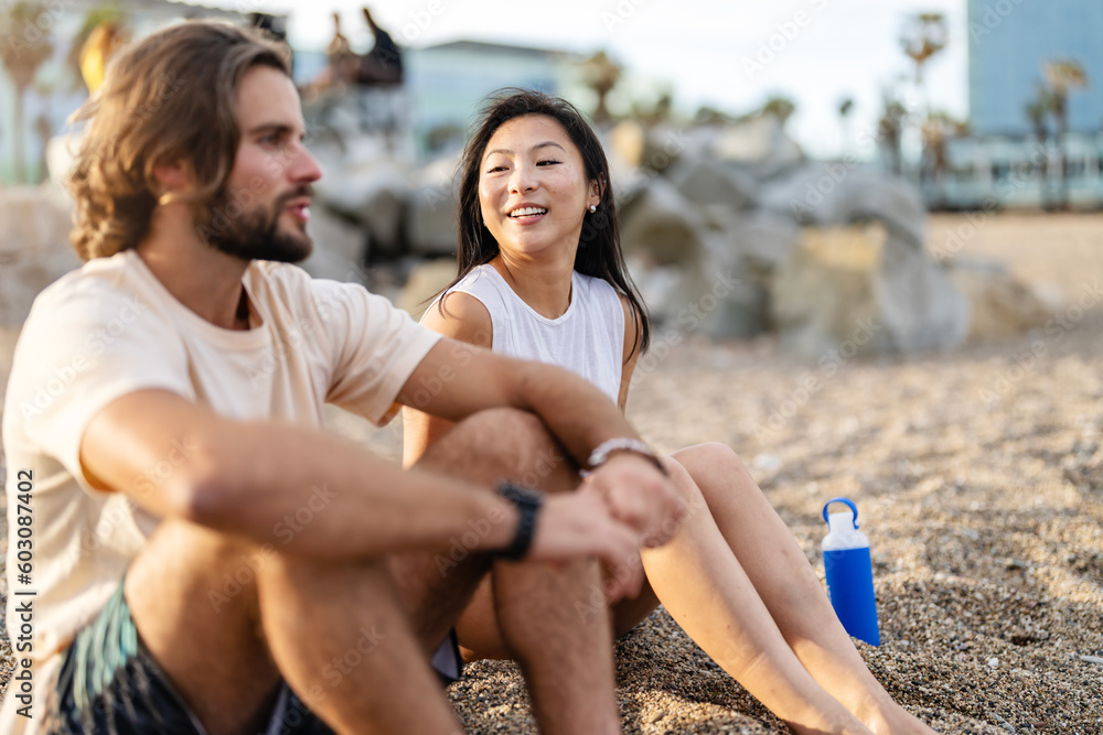 Young cheerful multi-ethnic couple sitting on the sandy sea beach in the sunset, talking Young cheerful multi-ethnic couple sitting on the sandy sea beach in the sunset, talking