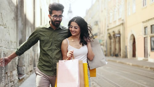 Cute young indian couple walking down with shopping bags on city street and talking outdoors Beautiful boyfriend and girlfriend enjoying great day with purchases after shopping together Sale Promotion
