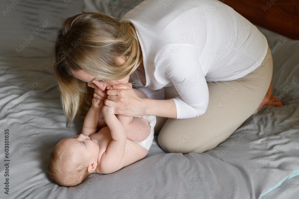 Joyful young mother plays with her baby lying on the bed,smiles,kisses,enjoys motherhood.Concept of family,love and care Joyful young mother plays with her baby lying on the bed,smiles,kisses,enjoys motherhood.Concept of family,love and care