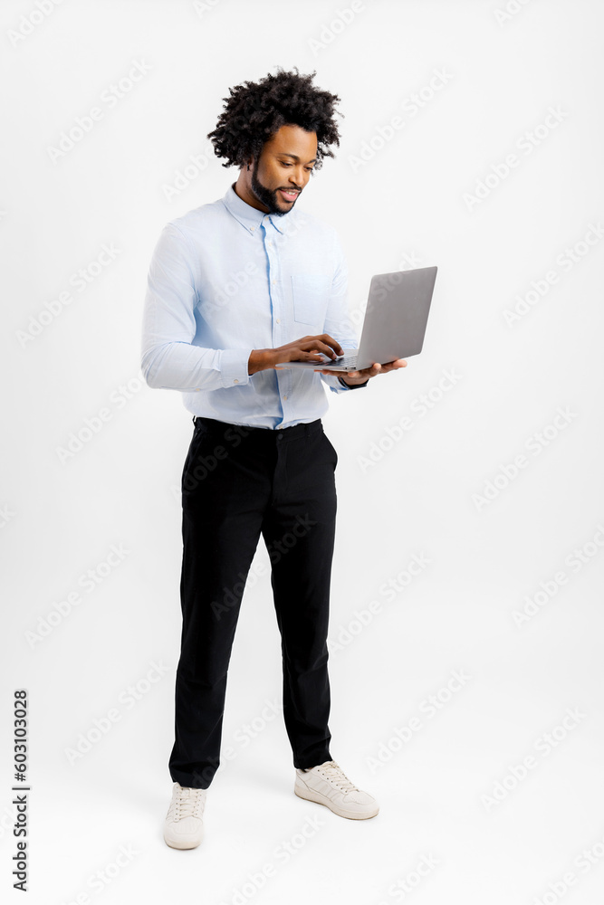 Full length portrait of curly young smiling african-american man using laptop computer isolated on white, freelancer guy typing, male office employee