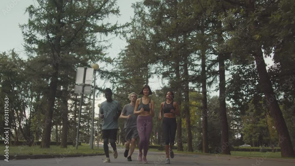 Low angle full length shot of four young diverse joggers jogging along track outdoors in green park moving towards camera Low angle full length shot of four young diverse joggers jogging along track outdoors in green park moving towards camera