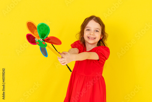 Photo of little cheerful girl in a red dress happy positive smile hold paper toy windmill isolated on yellow background