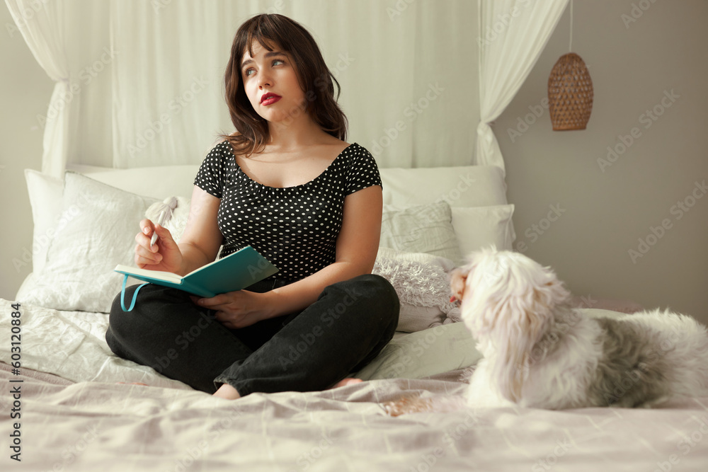 Female sitting on bed writing in diary while white chinese crested dog lying next to her, looking with trust and affection, girl planning next week, looking aside with thoughtful facial expression Female sitting on bed writing in diary while white chinese crested dog lying next to her, looking with trust and affection, girl planning next week, looking aside with thoughtful facial expression