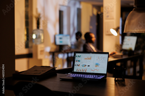 Selective focus on laptop computer with financial graph on screen, standing on desk table in startup office. In background employees working at marketing strategy late at night. Business concept