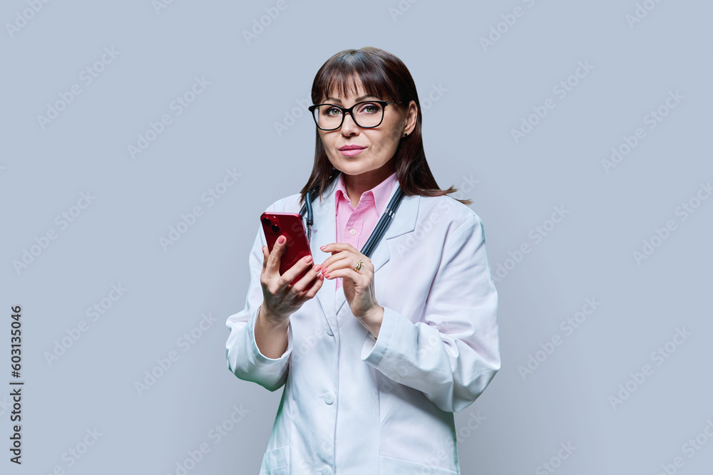 Mature female doctor in white lab coat using phone, on grey background Mature female doctor in white lab coat using phone, on grey background