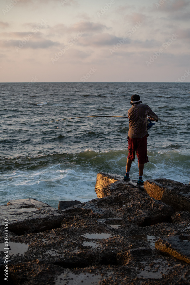 A man standing on rocks is fishing by the sea at sunset A man standing on rocks is fishing by the sea at sunset