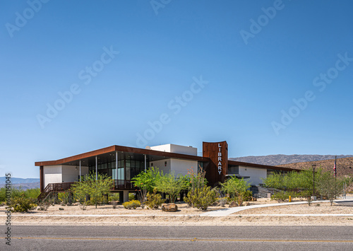 Borrego Springs, CA, USA - April 24, 2023: full frontal view on modern architecture style public Library building under blue sky, set on sandy desert floor with green bushes. US flag present