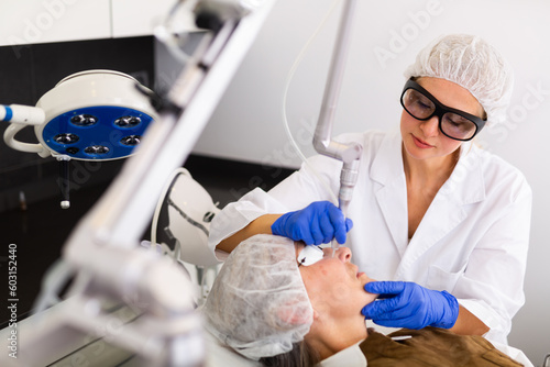 Cosmetologist woman in protective gloves and glasses doing laser wart removal procedure for elderly woman.