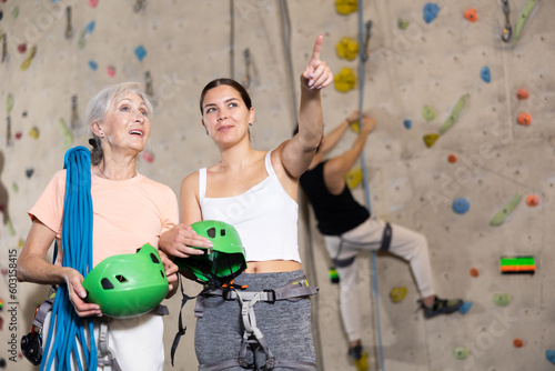 Young female trainer giving instructions to elderly woman about climbing artificial training rock wall in indoor bouldering gym