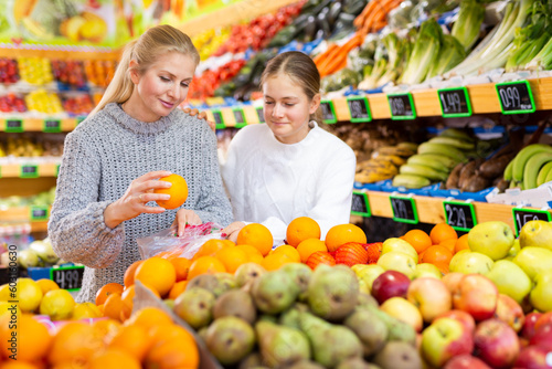 Cheerful teen girl with mother choosing sweet ripe oranges while shopping in greengrocery..